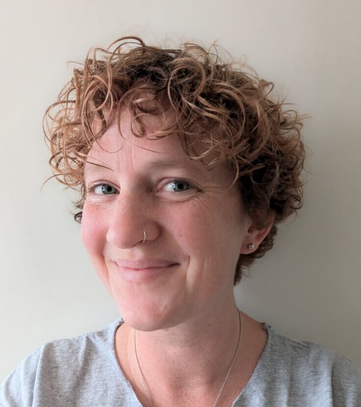 A photo of a white femme person with short curly hair and blue eyes. They are smiling at the camera. 